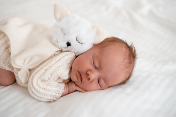 newborn baby sleeping on a white bed at home with a soft toy, close-up portrait of a newborn baby sleeping in a white knitted bodysuit, space and room for text