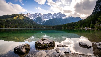 Scenic vista of a lake reflecting snow-capped mountains and a cloudy sky. Foreground features mossy rocks. Lush greenery flanks the water