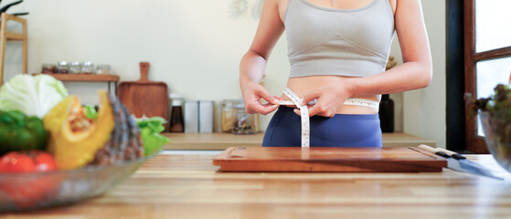 Happy asian woman measuring her waistline with tape measure while preparing healthy food in kitchen at home