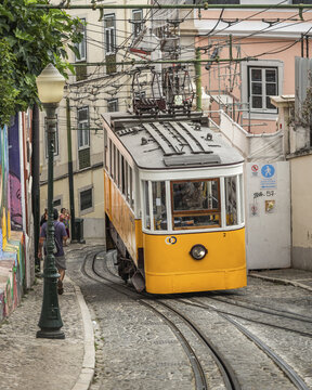 View of a vintage yellow tram ascends a steep cobblestone street, framed by colorful buildings and a classic lamppost, Lisbon, Lisbon, Portugal.