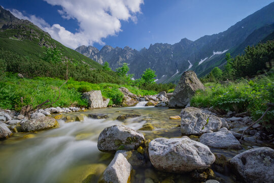 View of a pristine mountain stream flowing over smooth rocks, reflecting the vivid blue sky and lush green valley beneath towering peaks, Tatranska Javorina, Presovsky kraj, Slovakia. - Powered by Adobe