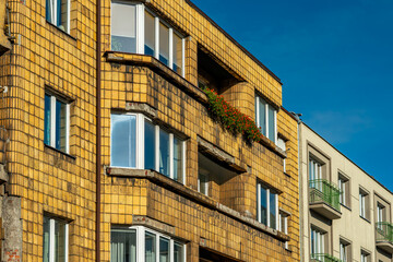 Old yellow residential building with distinctive tile facade, visible balconies and windows, showing signs of wear, contrasting with newer neighboring architecture.