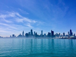 Obraz premium Dramatic panoramic view of the Chicago skyline from Lake Michigan, featuring modern skyscrapers like the John Hancock Center reflecting in serene blue water beneath a vast, clear summer sky