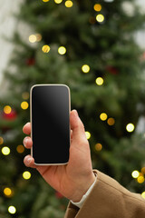 Man hand with smartphone with white blank screen against Christmas tree light garland and fairy lights. Festive Xmas Background. Mock up. Close up.