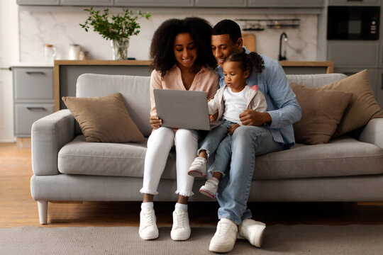 Cheerful black family having fun with laptop at home, watching photos or cartoons together, sitting on couch in living room, full length, copy space - Powered by Adobe