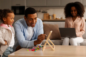 Empowering early education. Black father teaching little daughter alphabet with magnetic board while mother browsing internet on laptop, sitting on couch