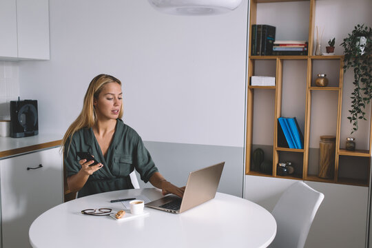 Concentrated young woman multitasking with smartphone and laptop while working remotely from home, maintaining a professional workflow at modern white table