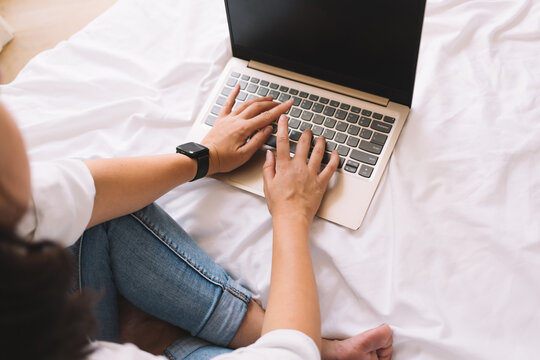 Close-up of woman typing on laptop while sitting cross-legged on bed, showcasing casual productivity and integration of smart tech into flexible digital routine