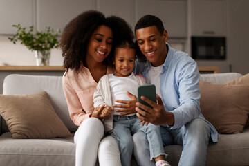 Happy African American parents and little kid using cellphone for video call or showing funny educational application, relaxing together on sofa at home