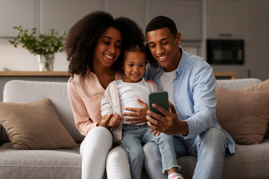 Happy African American parents and little kid using cellphone for video call or showing funny educational application, relaxing together on sofa at home - Powered by Adobe