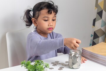 A little Asian girl putting coins into a glass jar. Learning to save money.