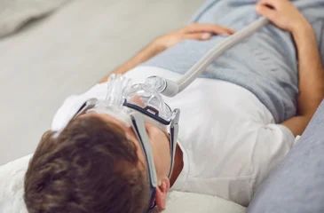 Fotobehang Leder Sick man with bronchitis, Covid 19 or asthma lying on couch with oxygen mask on face. Adult man with apnea disorder uses CPAP machine to help keep breathing airways open during sleep. Medical concept  © Studio Romantic