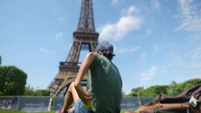 Person in cap and casual clothes lies on lush grass near sunglasses, holding smartphone with blurred eiffel tower in background, enjoying travel, technology and sunny park leisure