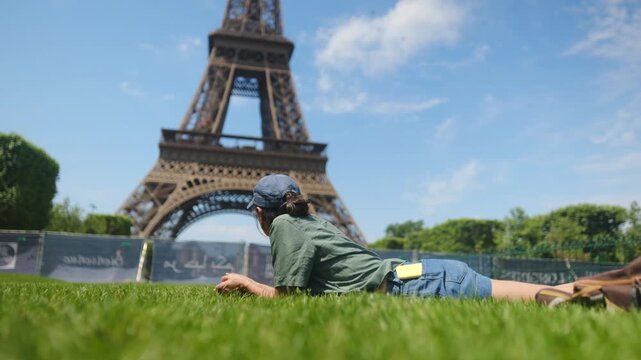 Person in cap and casual clothes lies on lush grass near sunglasses, holding smartphone with blurred eiffel tower in background, enjoying travel, technology and sunny park leisure