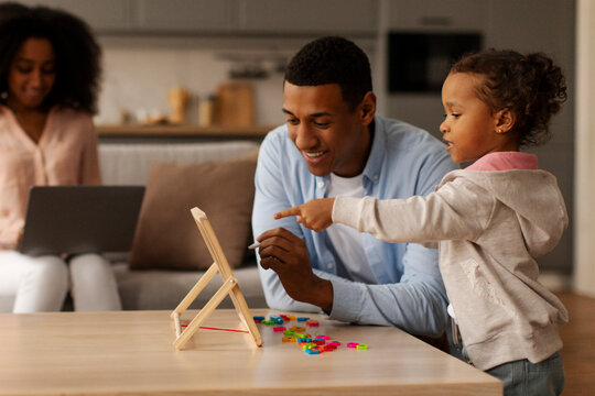 African American dad teaching little daughter and playing learning games with magnetic board while woman using laptop on background