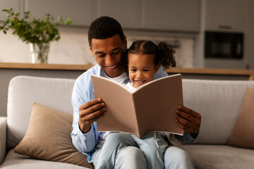 Loving African American father and his little daughter reading book together or looking photo album, girl sitting on daddy's laps on sofa in living room