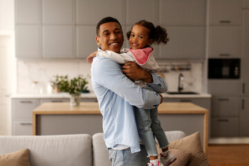 Happy childhood. Cute black little girl embracing his loving daddy and smiling together to camera, posing at home in living room interior