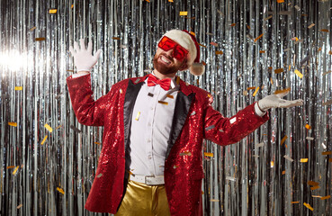 Festive Santa man in glitter costume, confetti. He raises hands with a smile before silver tinsel as flakes fall, capturing cheerful party celebration energy. Holiday Christmas and new year party.