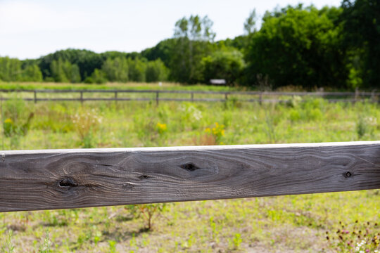 Close view of an old wood fence railing in the foreground with the other side of an overgrown riding pern for horses on a summer day in Maine.