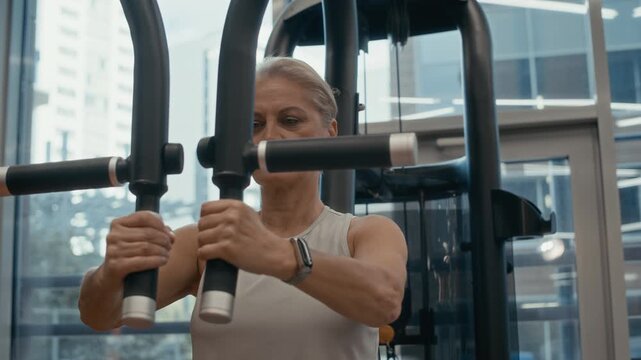Medium closeup shot of motivated senior Caucasian woman in sportswear building biceps while working out in modern well-equipped gym with floor-to-ceiling windows using chest press machine