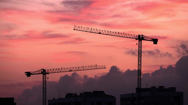 Silhouettes of construction cranes against a vibrant sunset sky. The scene captures the essence of urban development and industrial growth.