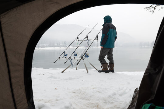 Fishing adventures. Camping and carp fishing with snow in winter on lake. Man near fishing gear covered in snow. Bad weather and cold conditions