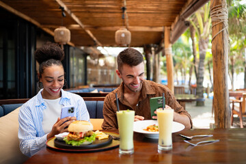 Diverse couple on a foodie date enjoys casual dining and a phone moment, sharing sunny vibes, drinks