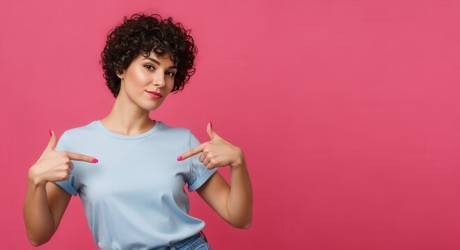 Confident young woman with curly hair pointing at herself. Female model wearing a blank blue t-shirt for a mockup on a pink background with copy space