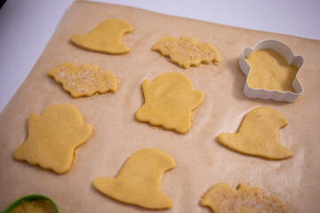 Close-up of a person preparing homemade cookies using Halloween-themed cutters. The hand sprinkles sugar on shaped dough pieces placed on parchment paper, ready for baking.