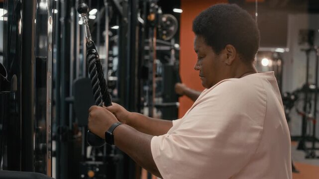 Side view slowmo shot of motivated plus size senior African American woman in sportswear building biceps while working out in modern gym using lat pulldown machine