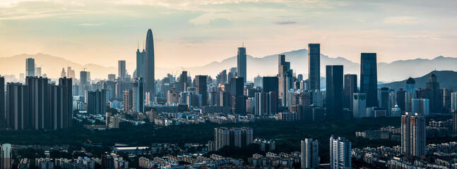 Telephoto view of the modern city skyline with skyscrapers silhouetted against a hazy mountain range at sunset in Shenzhen.