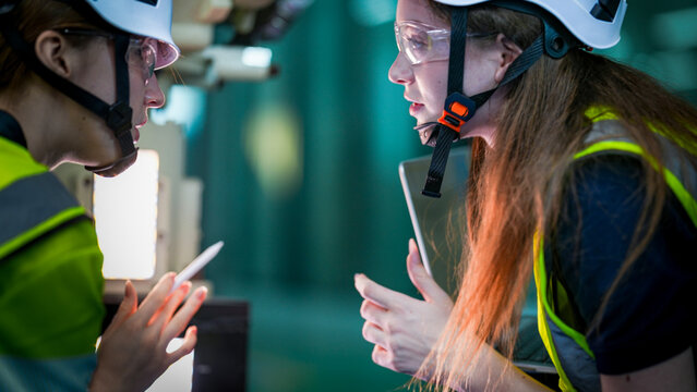 Two female robotics engineers in an R and D lab program an artificial intelligence cobot. They are testing the robotic system hardware and programming using a tablet.