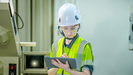 A female engineer in a hard hat and safety glasses inspects a high tech machine. She is holding a...