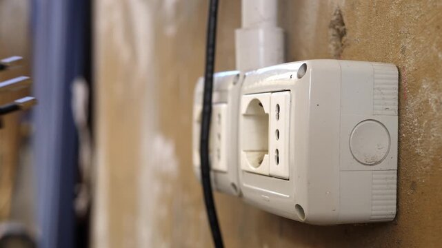 A woman's hand inserts an electrical plug into an outdoor wall socket fixed on a weathered wall, showing a simple, everyday action in a rustic setting.