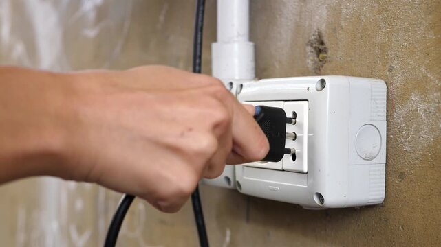 Close-up shot of a woman's hand pushing a plug into a worn-out exterior wall socket, emphasizing texture and tactile interaction.