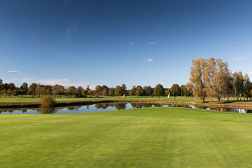 Fototapeta premium Golf green with flag and pond surrounded by autumn trees