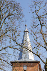 The tower of a Catholic church with a cross and a clock is photographed from below, amidst dry tree branches against a blue sky