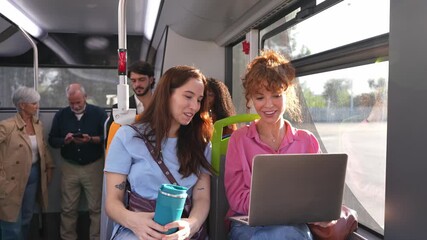 Two young women sitting together on a bus, working on a laptop. Diverse passengers commuting on public transportation in the city - Powered by Adobe