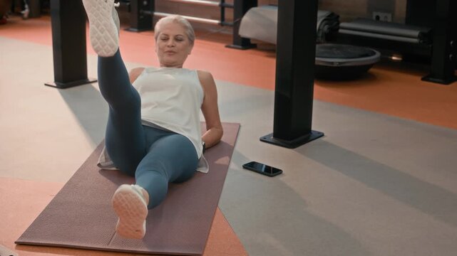 Slowmo shot of sporty senior Caucasian woman in activewear practicing lying leg lifts on fitness mat, exercising for better core strength and stability in modern gym