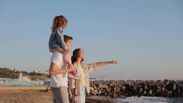 Side profile view young parents mom dad with little child kid daughter girl wear casual clothes point finer aside rest in free time walk on sea shore beach outdoor in summer day. Family day concept
