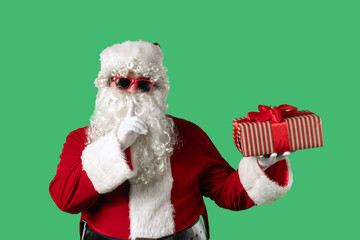 Santa Claus with a white curly beard, wearing a red suit and sunglasses, holds a gift box and makes a secret gesture on a green background. Ideal for Christmas-themed compositions and holiday layouts.