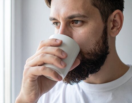 Close-up of a thoughtful bearded man enjoying a warm beverage from a white cup, finding a moment of peace
