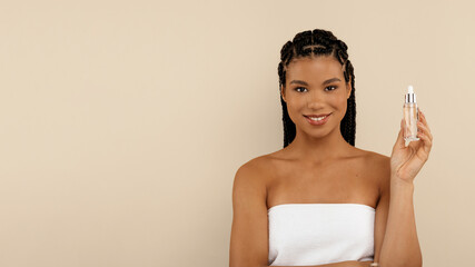 Smiling attractive good-looking half-naked millennial african woman wrapped in bath towel holding small bottle, recommending nourishing anti-age face serum or oil for young skin, beige studio