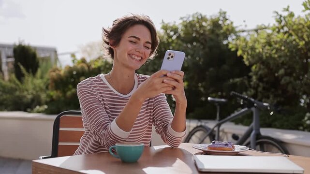 Young smiling happy woman she wears casual clothes sits at table in coffee shop cafe hold in hand use mobile cell phone relax rest in restaurant during free time outdoor. Freelance business concept