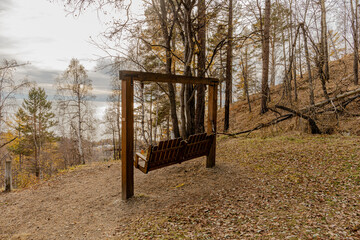 A wooden bench swing hangs from a sturdy frame in a sunlit autumn forest on sloped hill overlook autumn trees and a river or lake.  National Park at Baikal Lake