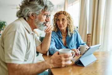 Portrait of a lovely tablet senior mature couple using a laptop together and having fun drinking coffee or tea sitting at a table at home