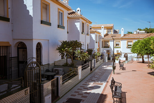 Traditional Andalusian residential street with whitewashed houses, tiled facades, potted plants and benches under bright sunlight, southern Spain architecture.