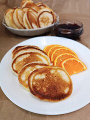 Delicious homemade pancakes served on a white plate with fresh orange slices and jam in the background. Tasty breakfast concept with golden pancakes, sweet sauce, and natural light