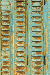 Old blue metal shutter with vents shows rust and tree shadows on a sunny day