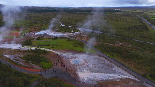 Aerial wide view shows steaming Haukadalur Geysir area in Iceland, blue pools, white terraces, red soil, green plain, visitors on boardwalks, roads, and tundra in overcast light.